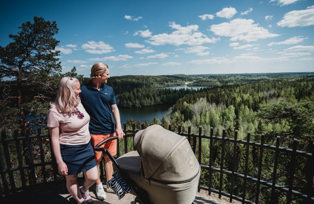 Pariskunta lastenvaunujen kanssa katselemassa maisemia näköalatasanteella. Näköalapaikalta avautuu kesäinen metsä- ja järvimaisema. Kuva: Maarit Vaahteranoksa