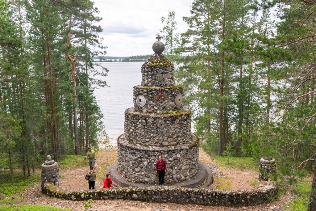 Neljä retkeilijää kivistä rakennetulla muistomerkillä. Taustalla on järvi.   For hikers are beside a monument which is built of stones. In the background there is a lake.