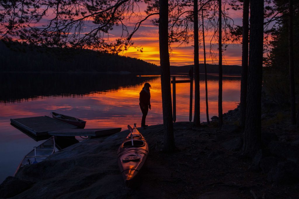 Kesäinen auringonlasku järvellä. Rannassa retkeilijä ja kajakki